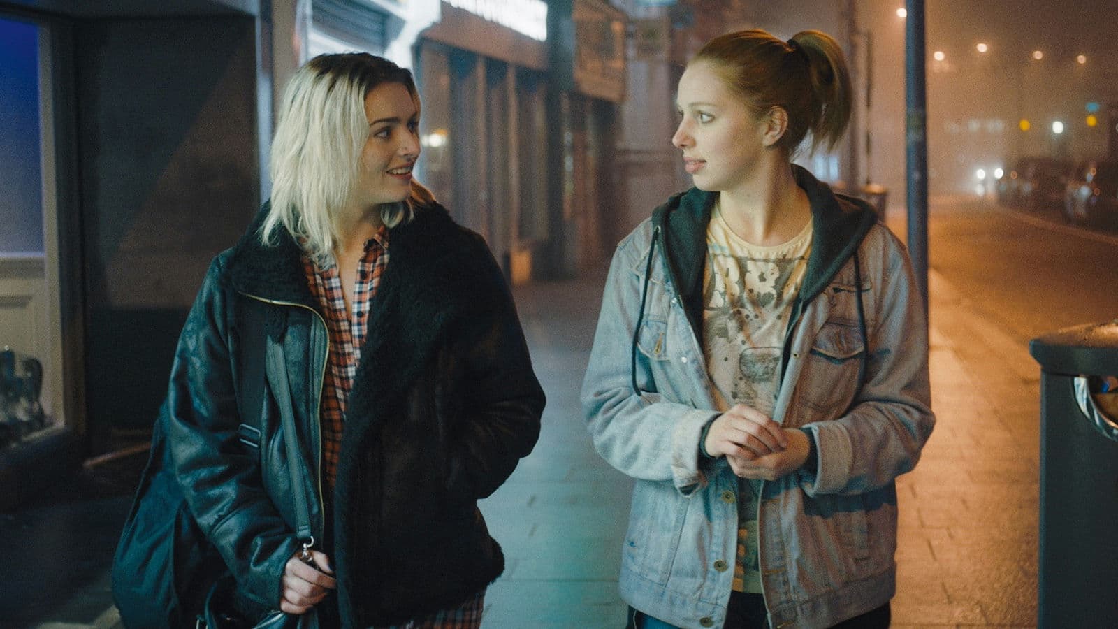 Two women are walking next to each other on a street, clearly talking and looking at each other.