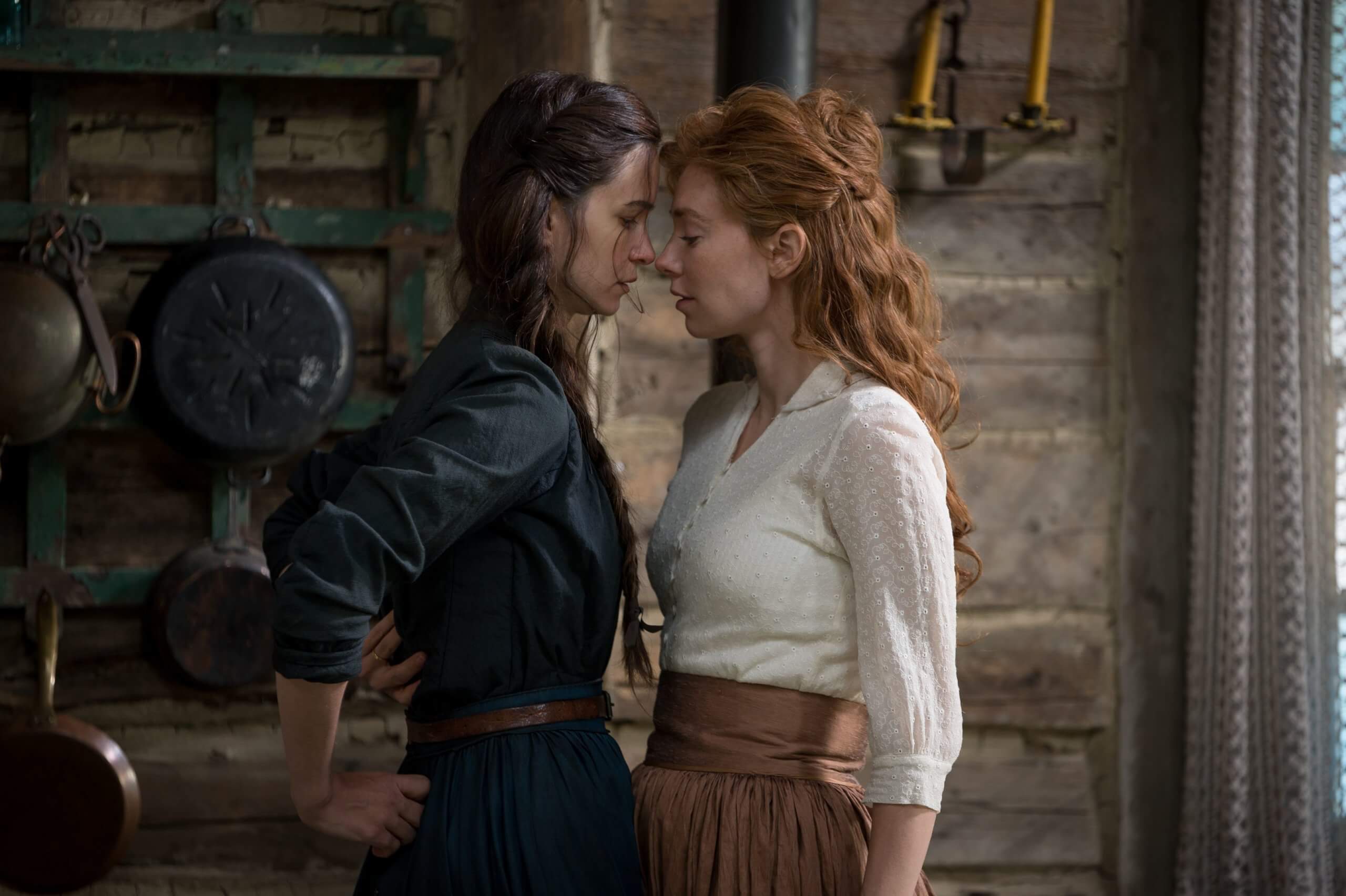 Two women with long hair are very close to each other, about to kiss. Their clothes and the background looks like 19th century, maybe earlier.