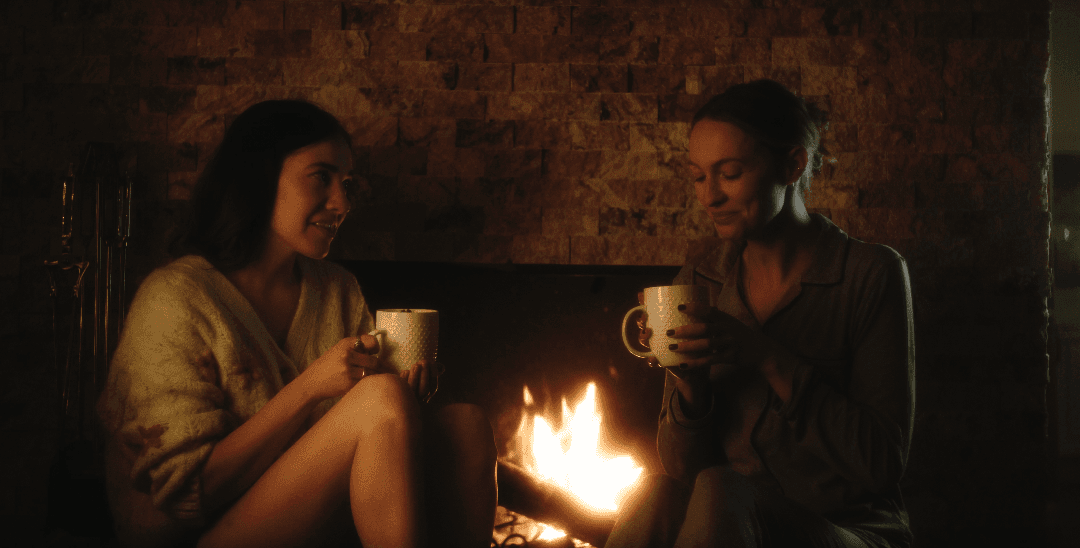 Two women sit beside a fireplace, drinking something from a mug.