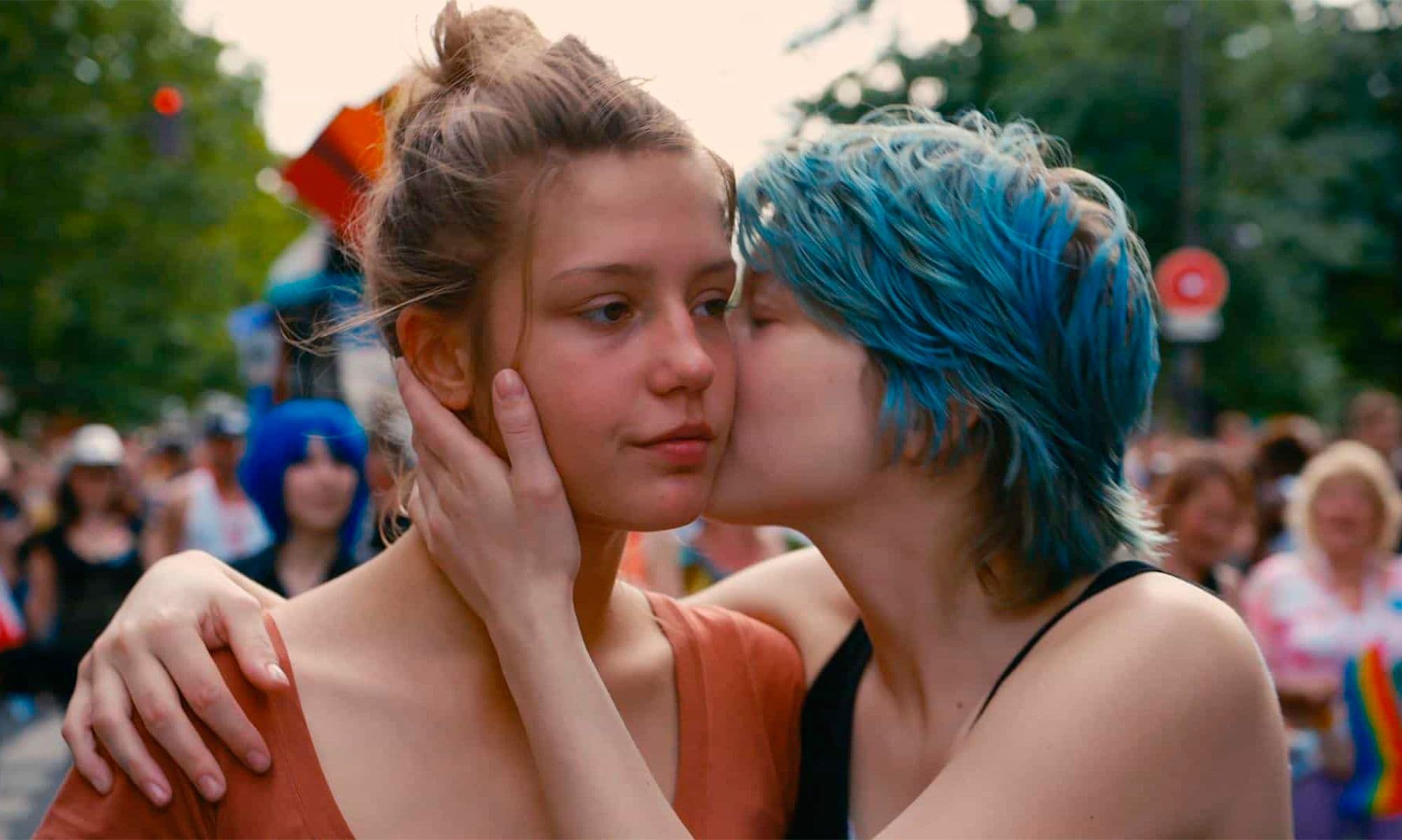 Two women are in a pride protests. One of them has short hair, dyied blue, and is kissing the other one in the cheek.