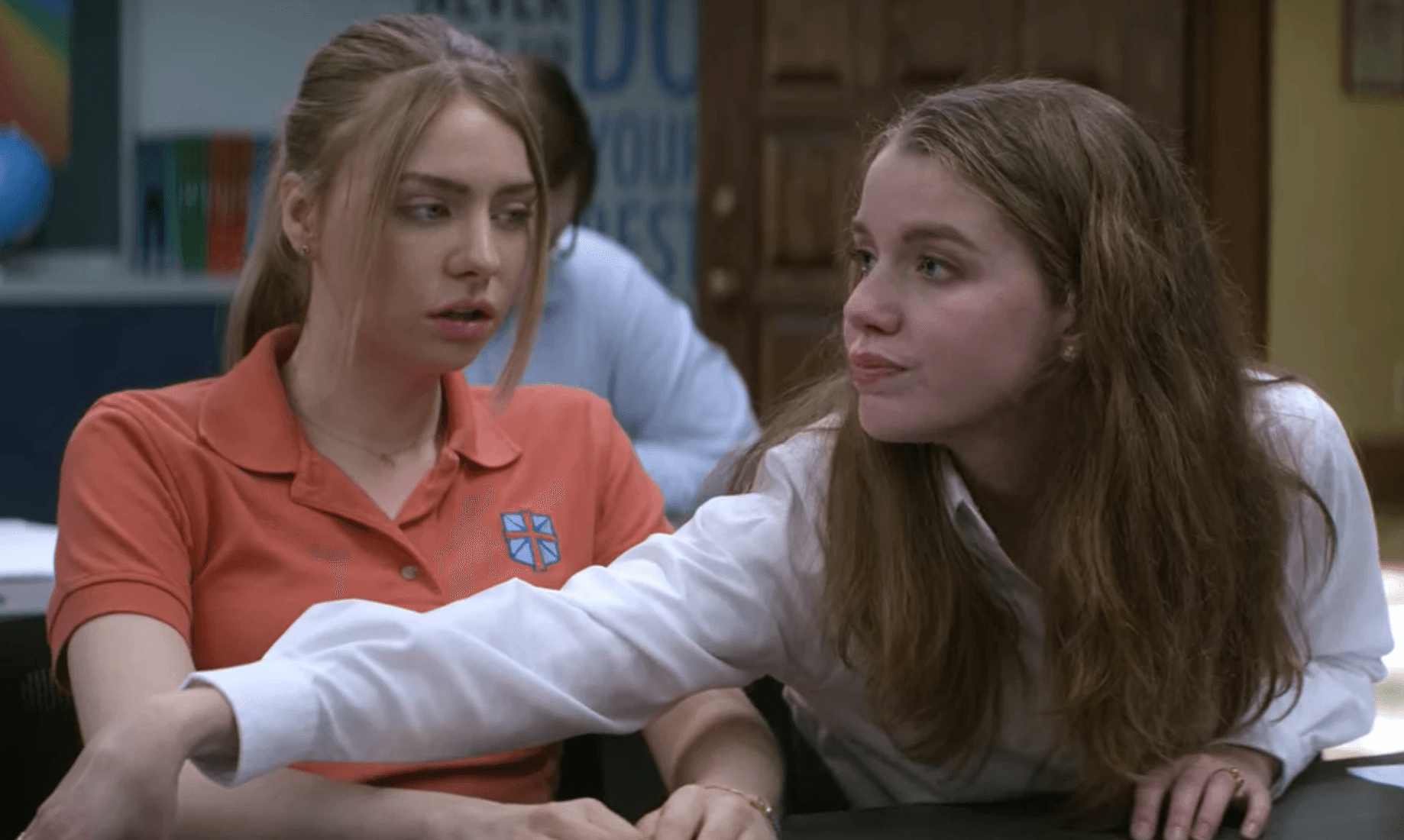 The image shows two young women in a classroom setting. One of them has her arm in front of the other's desk, and the other one is staring at her.