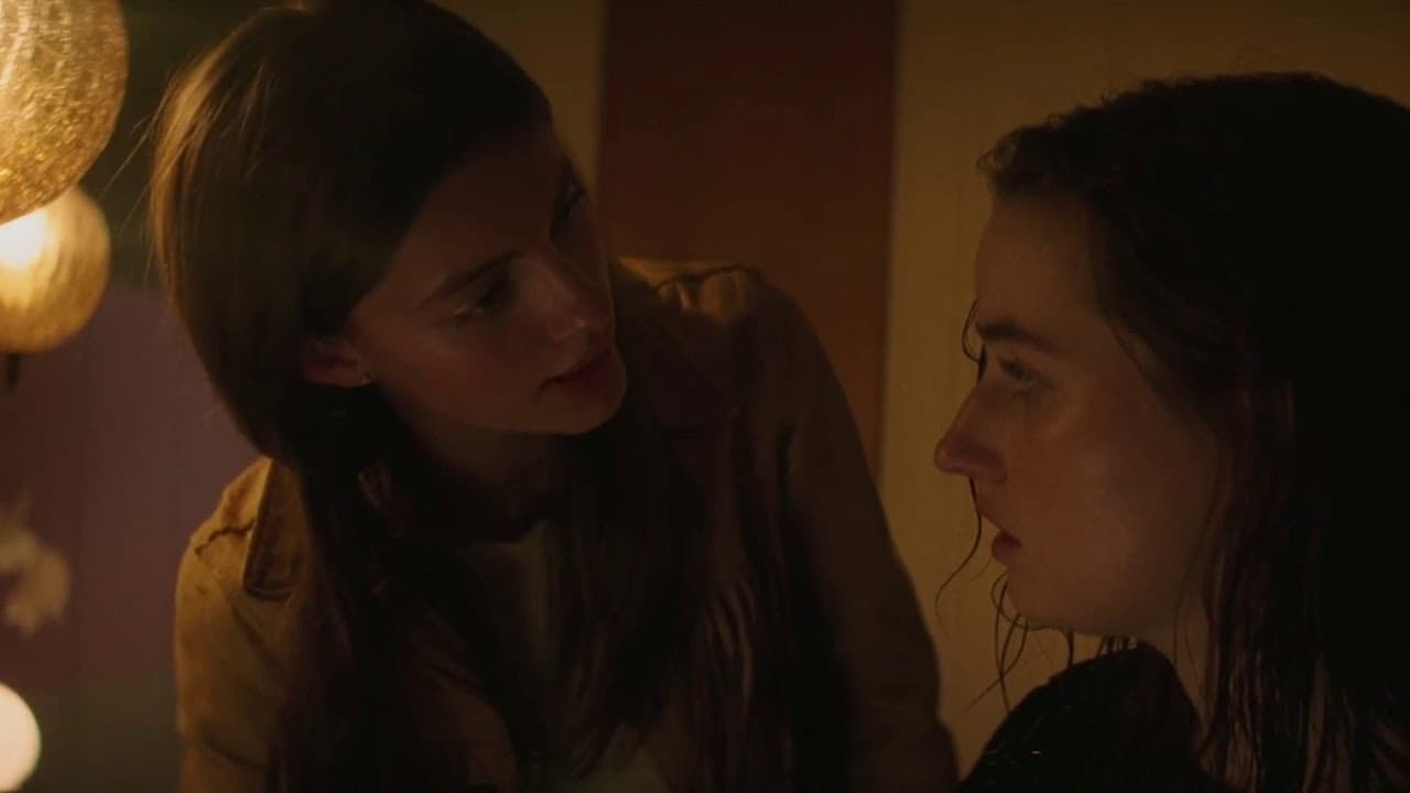 The image shows two young women, one with long dark hair and one with shorter brown hair, engaged in a close conversation. They appear to be looking intensely at each other.
