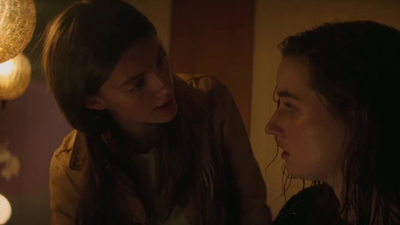 The image shows two young women, one with long dark hair and one with shorter brown hair, engaged in a close conversation. They appear to be looking intensely at each other.