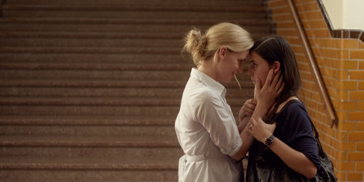 Frida and Mia are facing each other, their foreheads touching. Frida has her hands around Mia's neck.