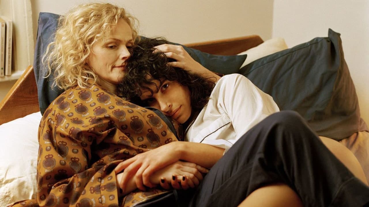 A blonde, curly-haired woman and a dark-curly-haired woman are lying on a bed, with the blonde comforting the other person.