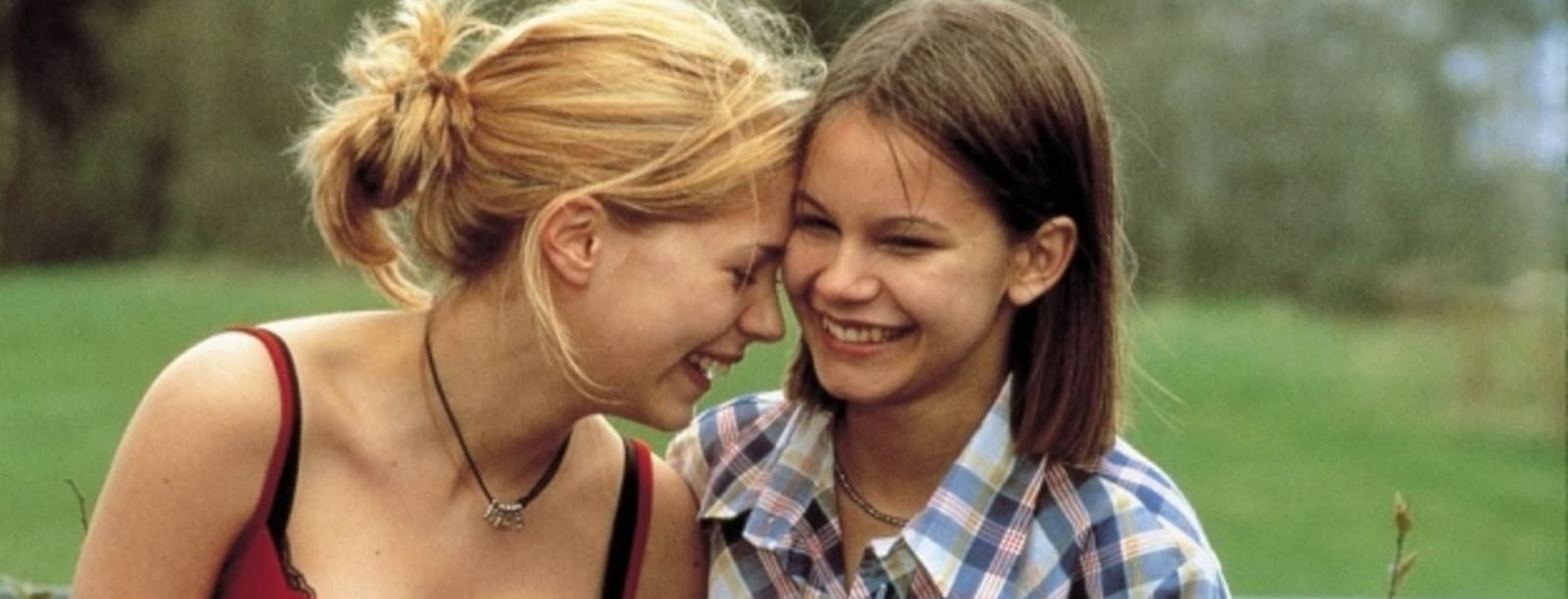 The image depicts two young women in an affectionate stance, their faces close together and expressions conveying a sense of joy and connection. The outdoor setting with the lush greenery creates a serene, natural backdrop for their tender interaction.