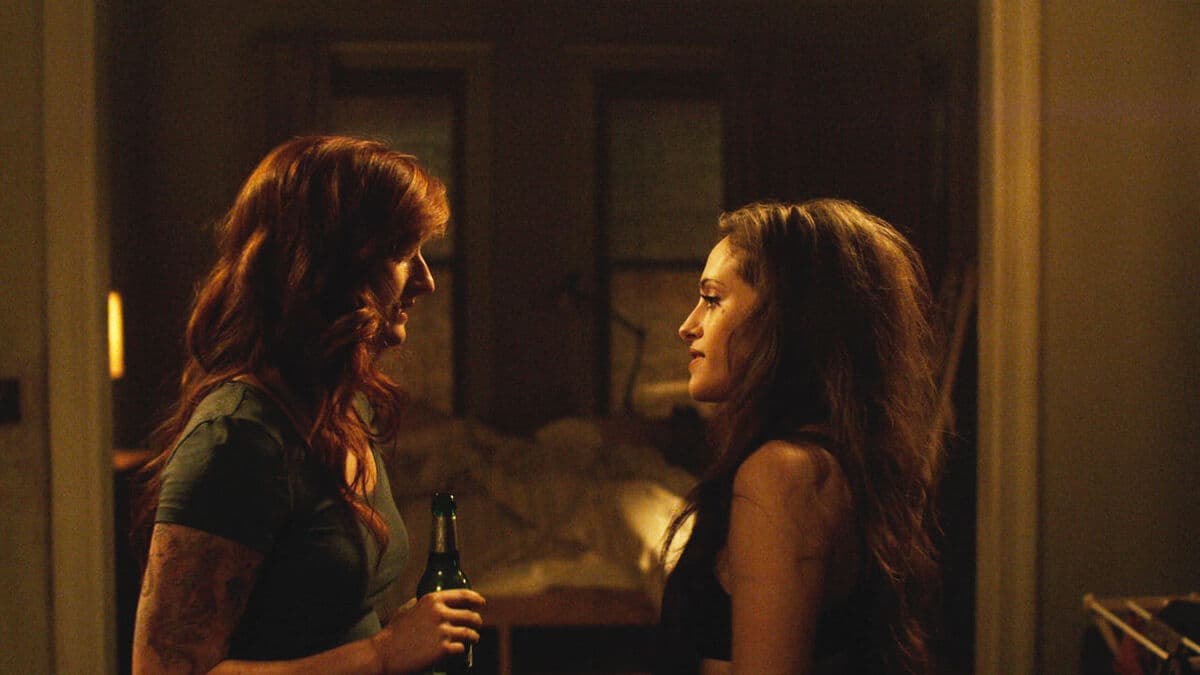 Two women facing each other in intimate conversation in a dimly lit room, one holding a bottle, with warm amber lighting creating a moody atmosphere.