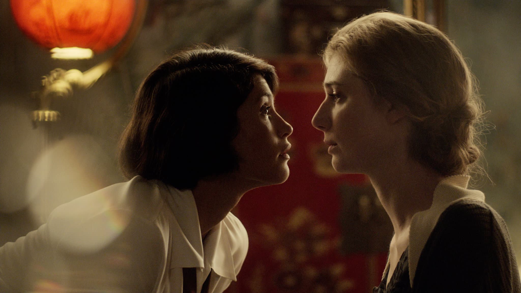 Two women face each other in an intimate close-up shot, their profiles silhouetted against a warm, atmospheric background with red lanterns. The lighting and composition create a sense of tension and yearning between them, with one woman leaning slightly forward toward the other.