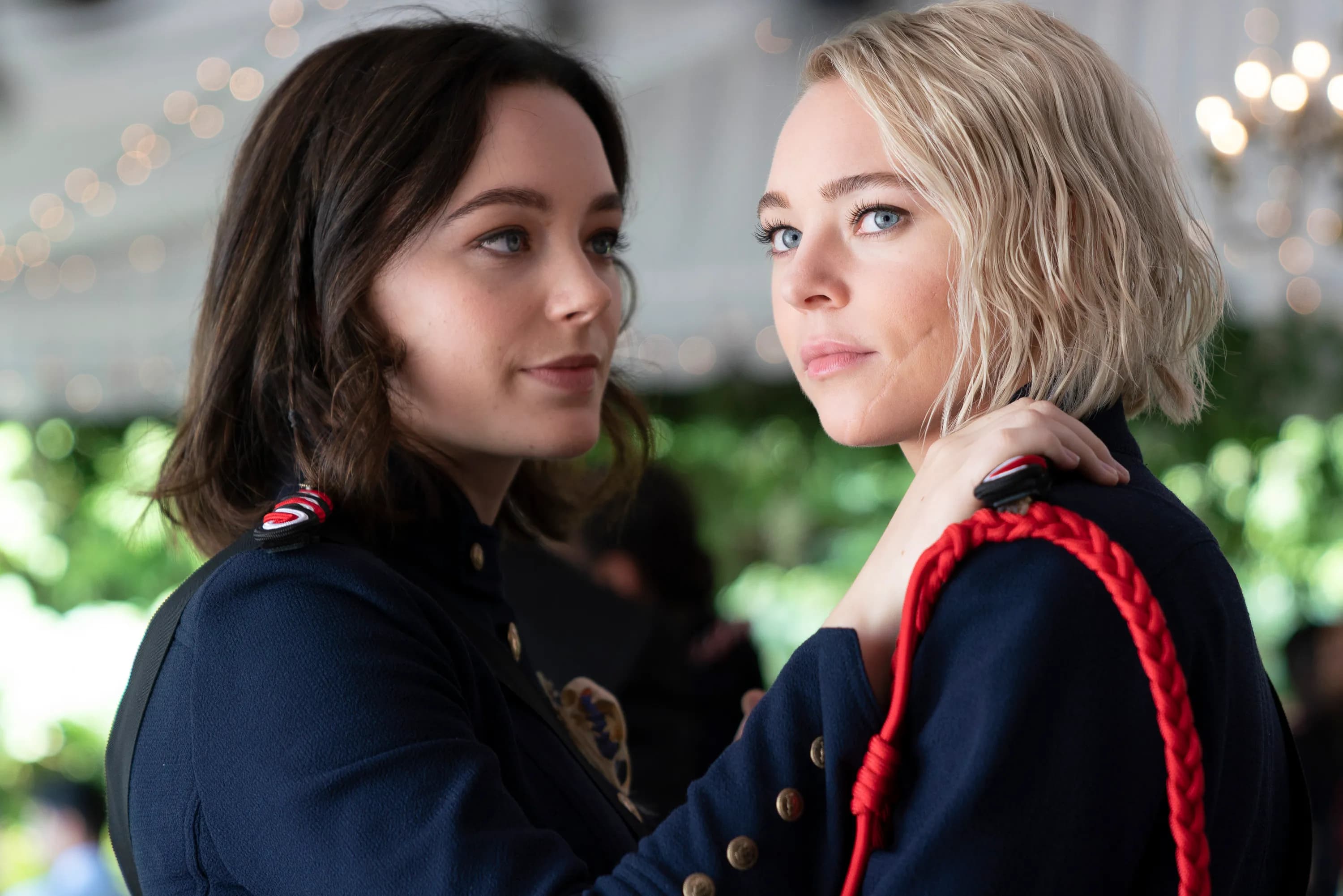 A blonde girl with braided hair and a brunette are very close to each other. They're both wearing a dark blue uniform, which looks military.