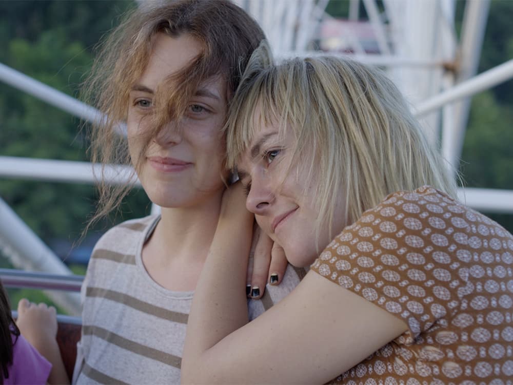 Two women are on a Ferris wheel, one of them has her head on the other's shoulder. They are both smiling.