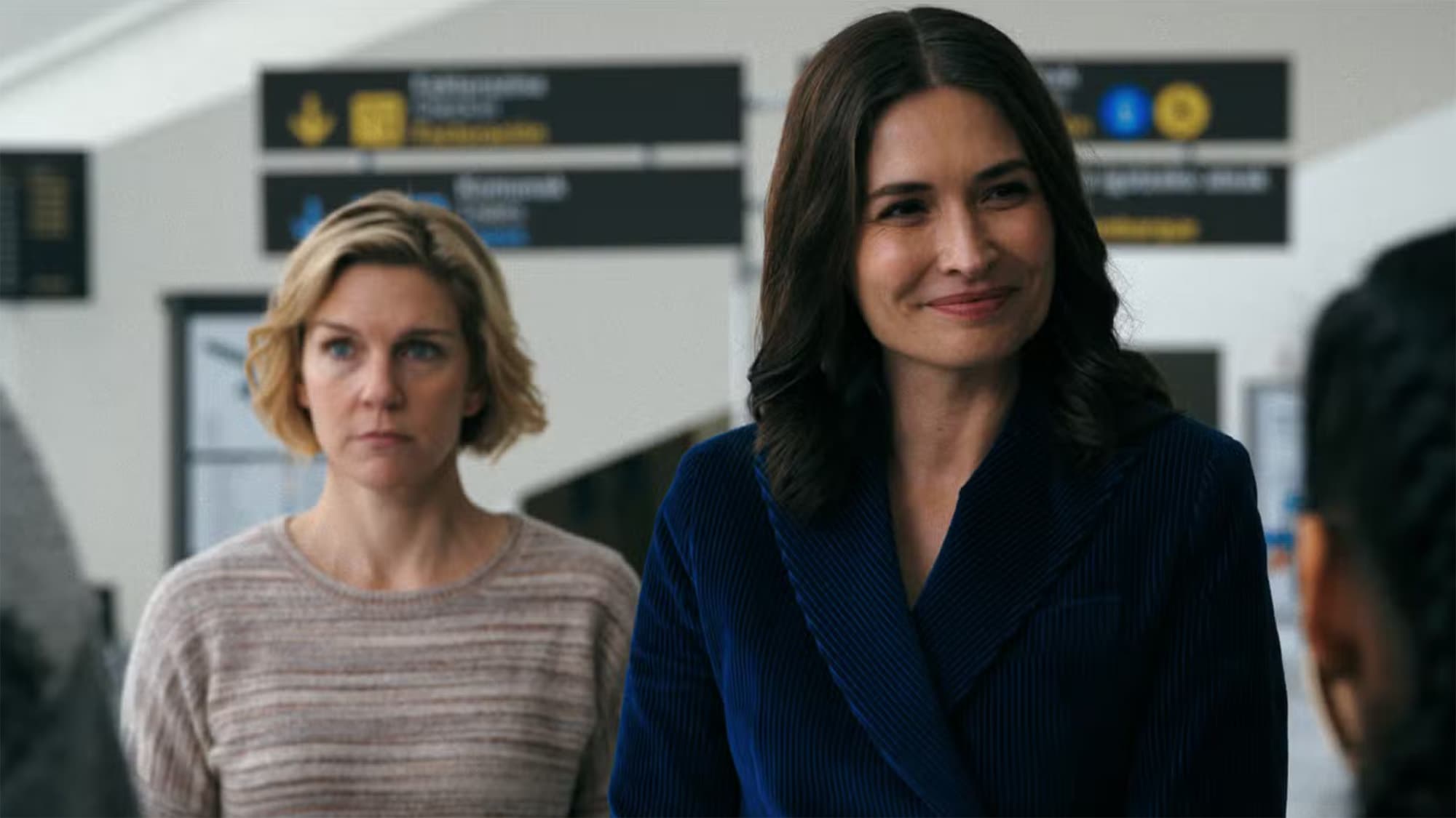Two women stand in an airport. One seems happy and put together and the other one seems in shock.